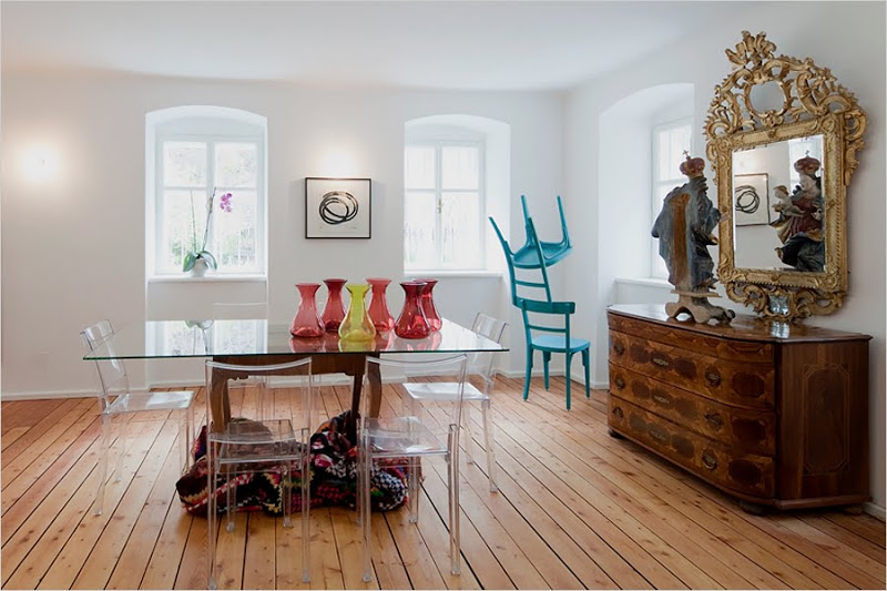 Dining room in the Hammerhaus castle with a knotty wood floor, Philippe Starck chairs, a custom dining table, a large antique mirror over a wooden chest of drawers and a turquoise chair sculpture by Markus Hofer
