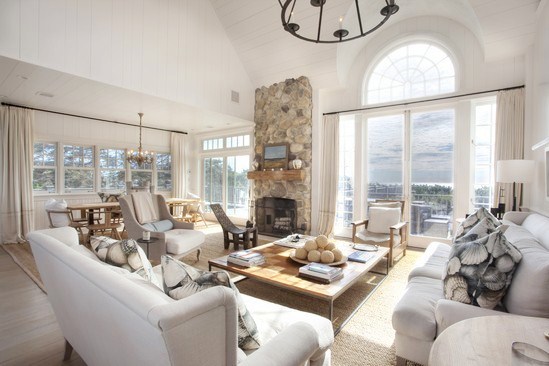 living room with french doors, neutral sofas and matching armchair with sea shell print accent pillows, a coffee table with a wooden top, fireplace with stone mantel, floor length curtains and a chandelier