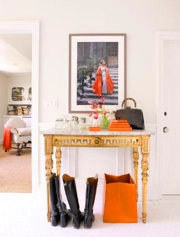 Foyer with an Hermes orange bag and two pairs of riding boots under a gilt table with marble top and a dramatic photo of woman in a ball gown descending a stair case