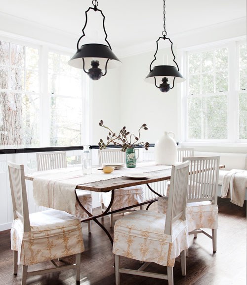 Breakfast nook with black pendant lights and a wood table surrounded by white chairs with upholstered seats