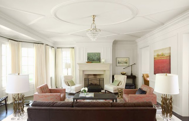 Living room with white moulded ceiling, a crystal ceiling medallion, a dark brown couch, patterned area rug, a dueling brick armchairs, a dark wood coffee table, a molded fireplace with two white armchairs next to it