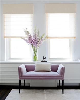 A lavender settee below two windows with a white accent pillow, on a white rug with dark wood floors
