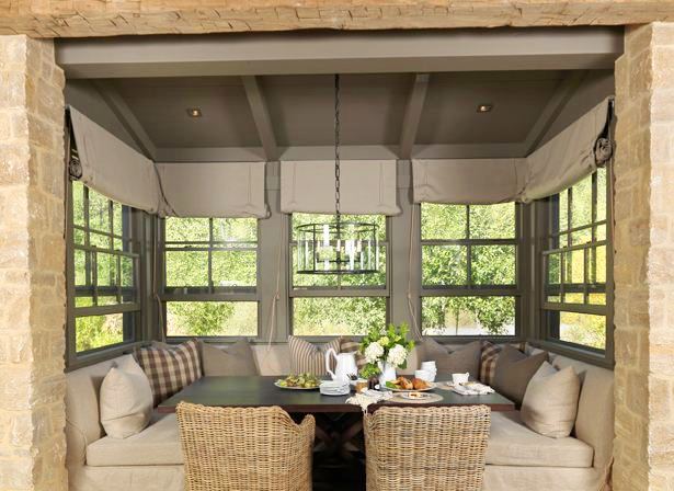 Rustic breakfast nook with built in banquet seating with two wicker chairs, casement windows with roll up shades, a dark wood table, a stone wall and a pendant light