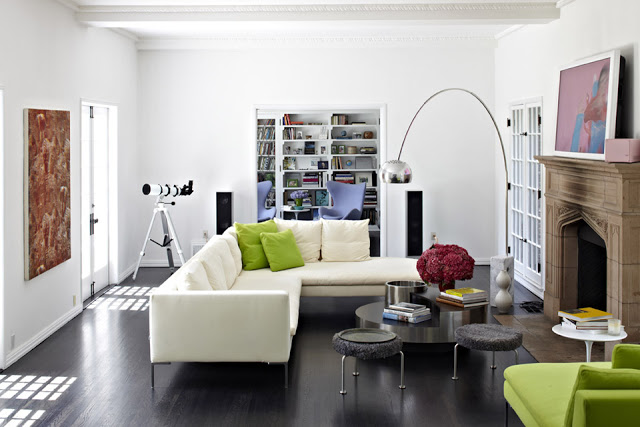 A modern living room with a silver 1962 Arco Lamp by Castiglioni, white sectional sofa, dark wood floor, a mirrored coffee table, a telescope, and a green armchair with a white round coffeetable