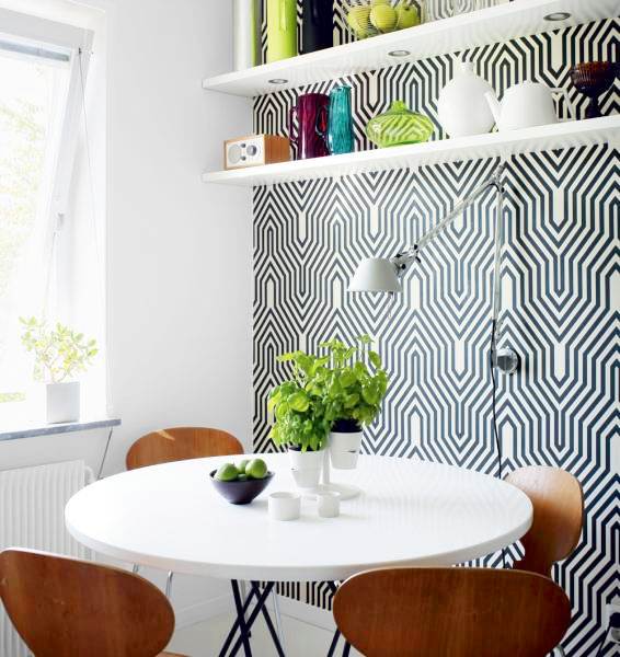 Breakfast nook with one wall covered in a bold graphic print, floating shelves holding bright colored containers, a white round table, and wooden Eames chairs