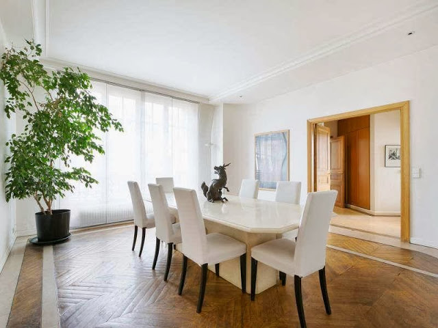 Dining room in a Paris apartment with herringbone wood floor, large windows, white high back chairs and a long hexagon table