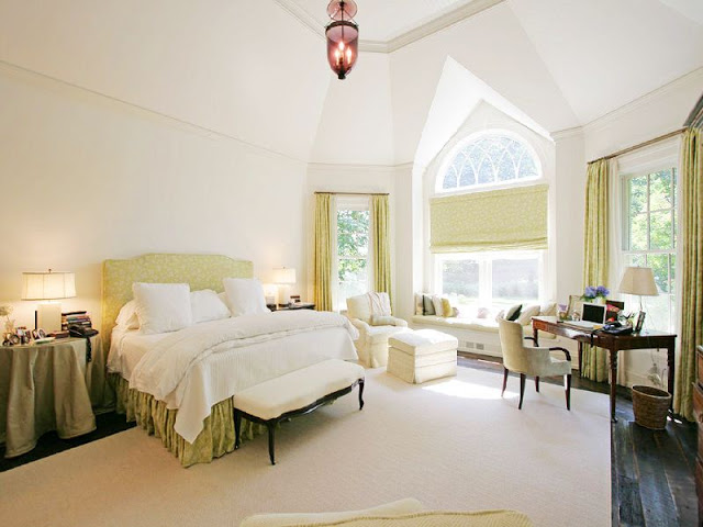 master bedroom with white walls and a high ceiling, a window seat, an green upholstered headboard with matching bed skirt, an ottoman at the foot of the bed and small home office with a rustic wood floor