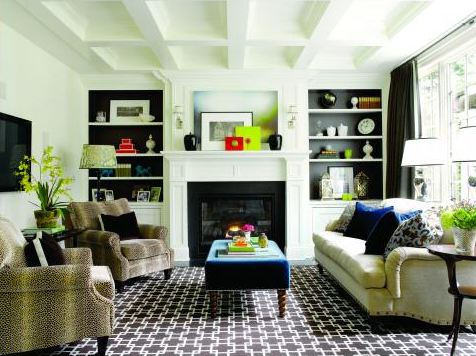 Family room designed by Graciela Rutkowski with trellis patterned carpet, two animal print armchairs, a taupe sofa with blue accent pillows, a blue upholstered ottoman, a black fireplace with a white frame, built in bookcases with the back of the shelves painted black and a coffered ceiling 
