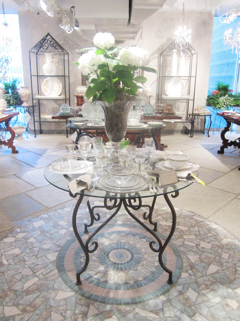 Round glass table with metal legs on a mosaic floor with a crystal table setting and a large white hydrangea plant in a glass vase
