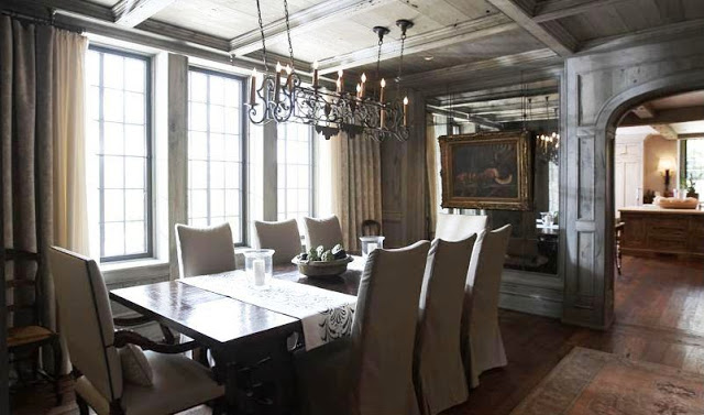 Formal dining room with grey wash wood paneling with exposed beams, encasement windows, neutral floor length curtains, wood floor, a long dark wood table surrounded by upholstered high back chairs and a wire chandelier