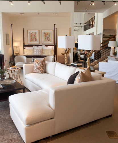 White L-shaped couch with accent pillows with an accent table holding two lamps with gold sculpted bases and white shades, in the background a large bed with a dark wood canopy frame has a COCOCOZY pillow on it