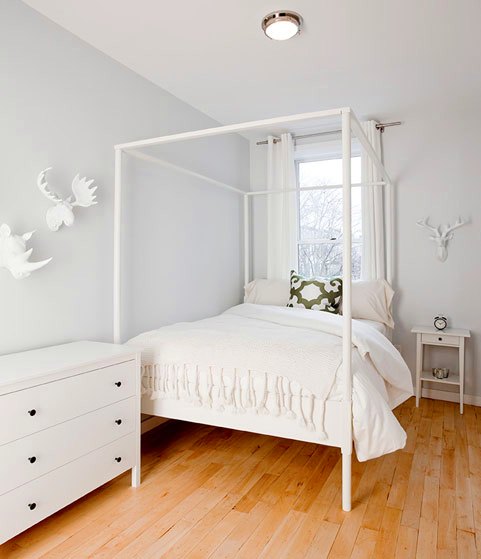 White bedroom with a chest of drawers, canopy bed, and white animal head sculpture, wood floor and a green and white accent pillow