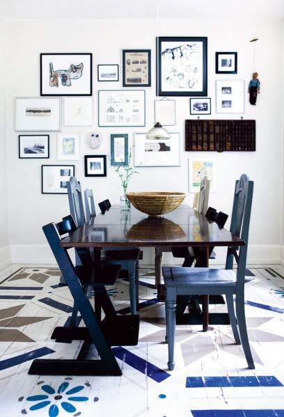 Dining room with dark wood table surrounded by mismatched blue chairs, the white wall in the back is covered with art in different sizes in blue frames, and a single pendant light. The white wood floor is painted with a variety of blue, brown and grey graphic patterns