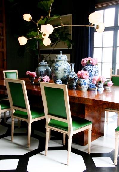 Dark dining room with black and white painted floor, brown stone table surrounded by upholstered green chairs, on the table are blue and white chinese pots in different sizes and shapes and above it all if a modern chandelier