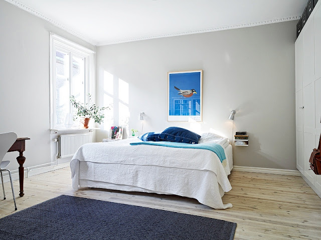 bedroom in a tiny apartment with blue rug, light wood floors, grey walls, a floating nightstand with wall mounted lights