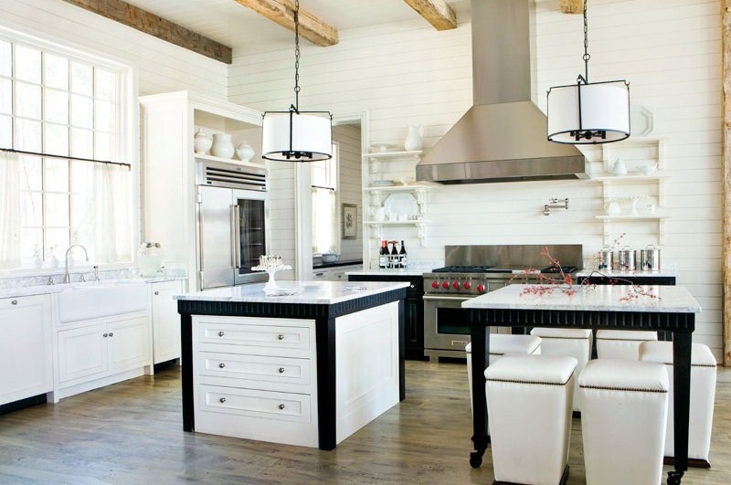White rustic kitchen with stainless appliances, wood floor, two black and white pendant lights, Carrara marble countertops, and two islands one for preparing food which has white drawers and black detailing and the other for dining which is surrounded by white stools