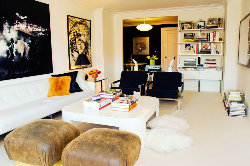 Living room with a white tufted sofa with gold and black accent pillows, leather ottomans, a white coffee table, black armchairs, built in bookshelves and modern art on the walls