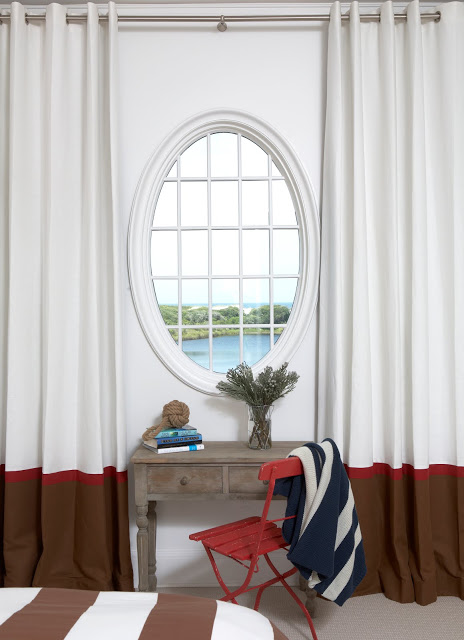 desk in a beach house with an oval mirror, white drapes with a single brown and red stripe on the bottom with a red metal beach chair