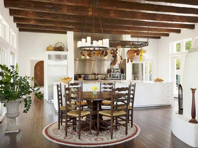 Open kitchen with round wood breakfast table surrounded by high back wood chairs on a round rug, wood floors, exposed beams and a metal chandelier