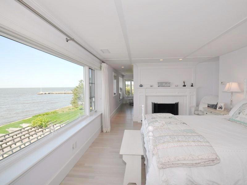 White master bedroomwith a light wood floor, bench style ottoman at the foot of the bed, a fireplace and a picture window with a view of the Long Island Song