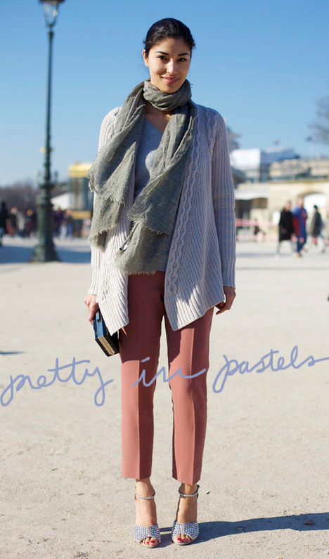 Woman wearing coral ankle length pants, a chunky cream sweater with a pale green woven scarf. She's holding a black clutch purse with silver clasp and strapy, peep toe metallic heels