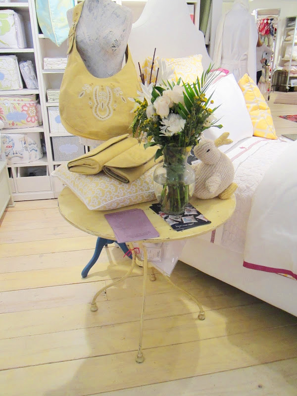 Close up of the night table in Serena & Lily's bright vignette. The night stand is round and yellow with wire legs and it holds a glass jug of flowers, a stuffed rabbit, two yellow purses and a yellow and white pillow. 