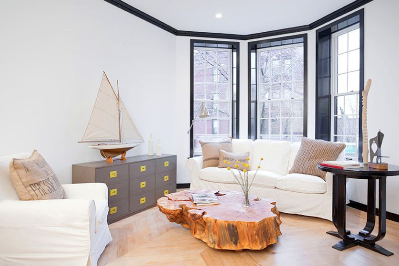 Living room with black trimmed windows, a white sofa and matching armchair, herringbone wood floor, tree trunk coffee table, grey chest of drawers with yellow drawer pulls and a ship on the top and a black table