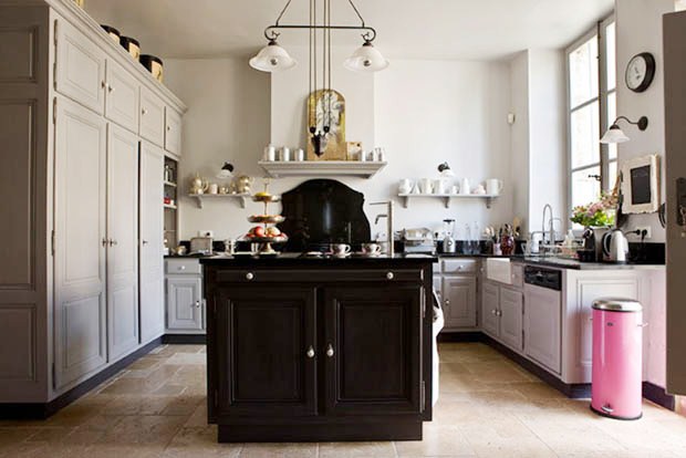 Kitchen in a french mansion with a black island, grey cabinets and drawers, a chandelier, black counter tops, a stone tile floor and a pink trash can