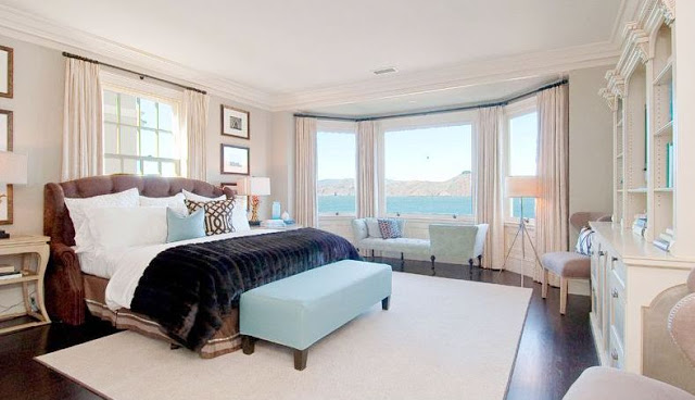 Master Suite with ocean view, dark wood floor, upholstered headboard, floor length curtains, white rug and a light blue ottoman
