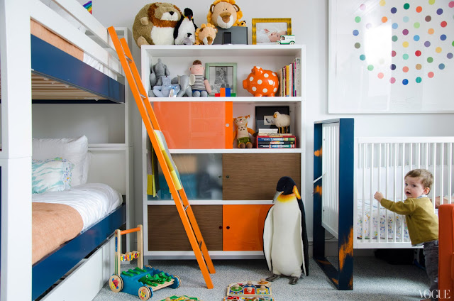 Kid's bedroom with white shelves with orange and brown doors, bunk beds and a crib in matching blue and white frame, orange accents on the ladder and crib and a large framed picture of a circle with colored dots arranged in a circle inside it