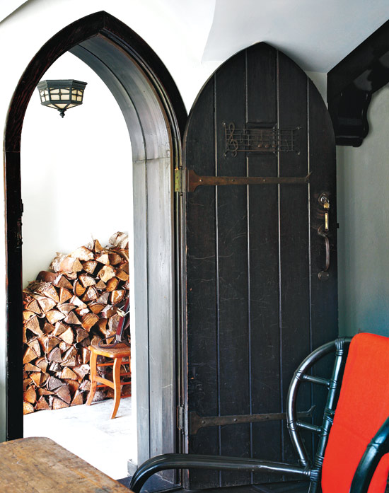 Alternative view of the foyer showing it's dark wood arched door, a dark wood chair with a red blanket draped over the back, and a stack of firewood creating a visually interesting and functional feature wall