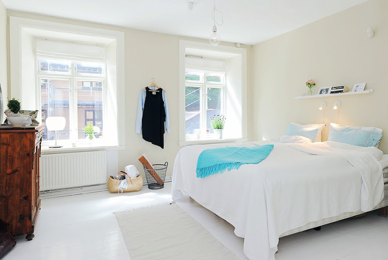 White minimalist bedroom with turquoise throw, blue and white patterned pillows, sea grass rug at the foot of the bed and wood chest of drawers