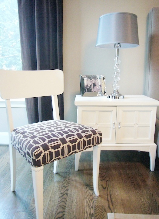 The side chair painted bright white and upholstered with Kelly Wearstler's brown Emerald Cut fabric on the seat in a bedroom with a hardwood floor, grey curtains, white nightstand with a framed photo and a lamp with a glass base