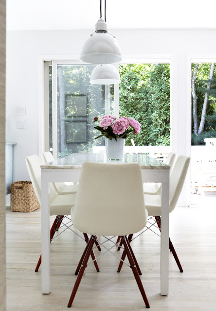 Dining room with white table with a marble top holding a vase of pink flowers surrounded by white chairs with wooden legs overlooking a lush garden