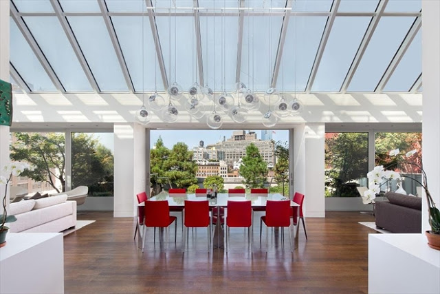 Penthouse in Tribeca with a glass ceiling and large glass windows overlooking a cityscape. A white table is surrounded by red chairs is placed facing the central window