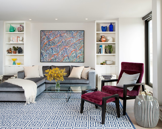 Modern living room with graphic blue Greek key rug, silver blue velvet sectional sofa, glass coffee table and burgundy accent chair and ottoman