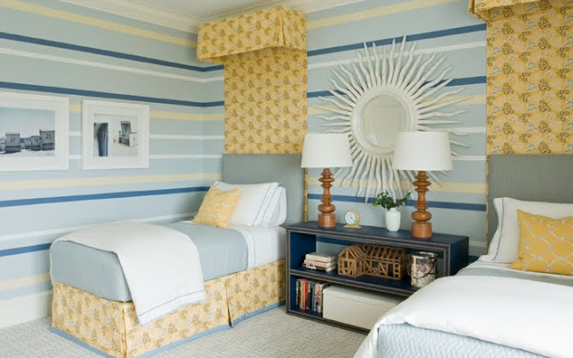 Striped walls in shades of blue and yellow coordinate with simple blue upholstered headbaords with a yellow floral crown and drape that match the pleated bedskirts in a twin bedroom