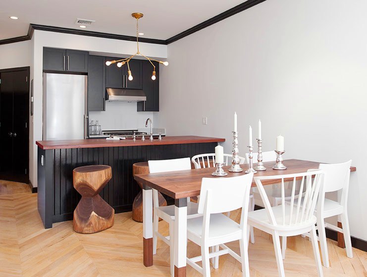 Kitchen and dining area with black cabinets and drawers, stainless appliance, herringbone wood floor, a branch inspired chandelier, a wood table, mismatched painted white wood hairs and carved stools made from tree trunks