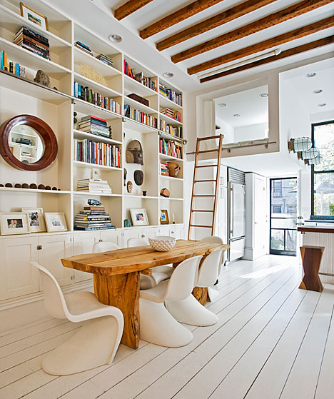 Living room in a home in NYC with wide plank white wood floors, built in bookshelves, a reclaimed wood table surrounded by white S Chairs and exposed beams