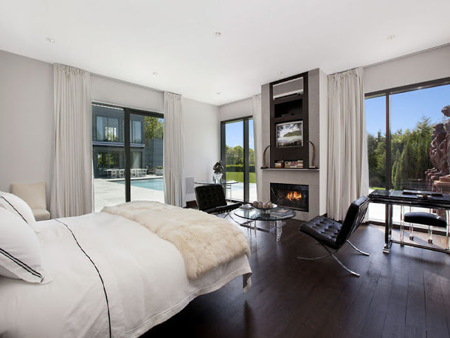 Bedroom with white bed, surrounded by large windows with floor length white curtains overlooking the backyard with a pool