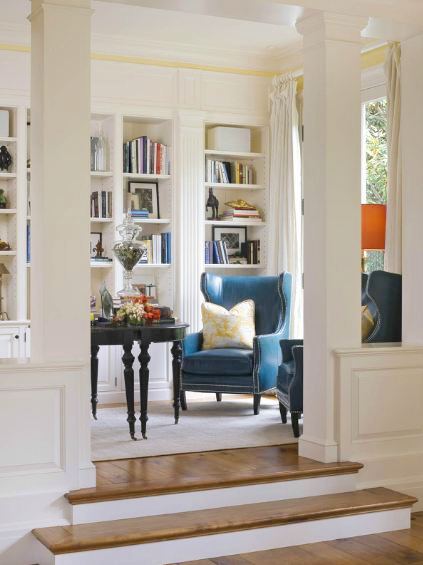 Library in a san francisco mansion with two blue wingback chairs with nail head trim, a black table, wide paneled wood floor, white built in book shelves, white floor length curtains and a white area rug