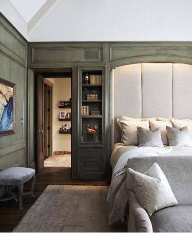 Master bedroom with green stained wood paneling and built-in shelving surround an oversized upholstered headboard, at the foot of the bed is a grey sofa with matching accent pillow, the room has dark wood flooring and a large neutral area rug