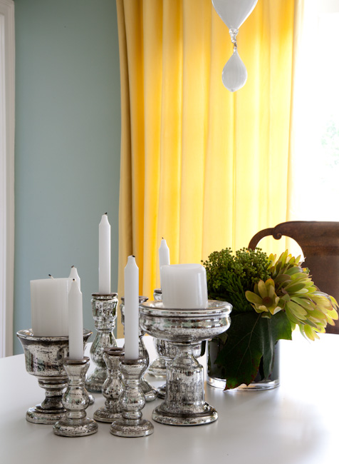 Teresa's Green paint in a dining room with yellow curtains, a white table and wood chairs. On the table is an assortment of mercury candle stick holders