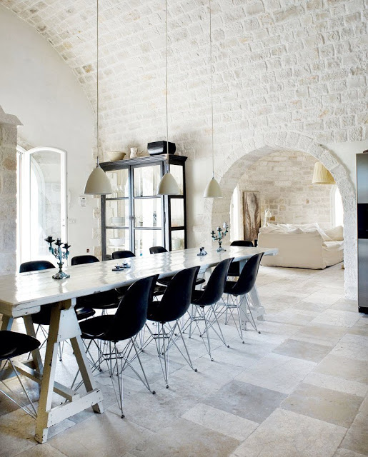 black Eames chairs surrounding a long table in a white dining room with white pendant lights, a long table and painted brick wall with arched entryway into a living room