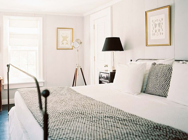 White bedroom with paneled walls, a dark wood floor, iron bed frame, black pillow with white poka dots and a matching throw pillow at the foot of the bed. The black nightstand has a tall brass lamp with a black lampshade