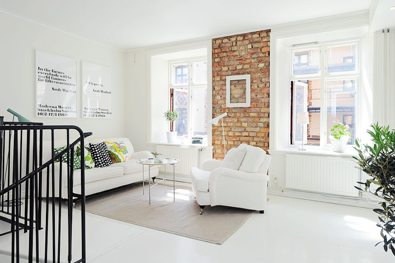 Living room with painted white wood floor, round cofee table, exposed brick wall with an empty white picture frame, a white sofa with bright patterned accent pillows, a white armchair and a taupe rug