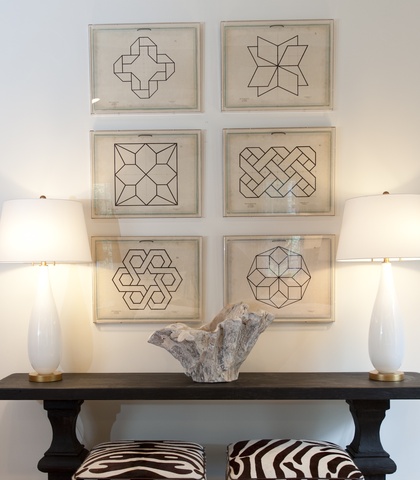 a long black accent table with two white lamps on each end and two zebra print stools sit in front of a white wall with six pictures of geometric shapes stacked on top of each other in two columns 