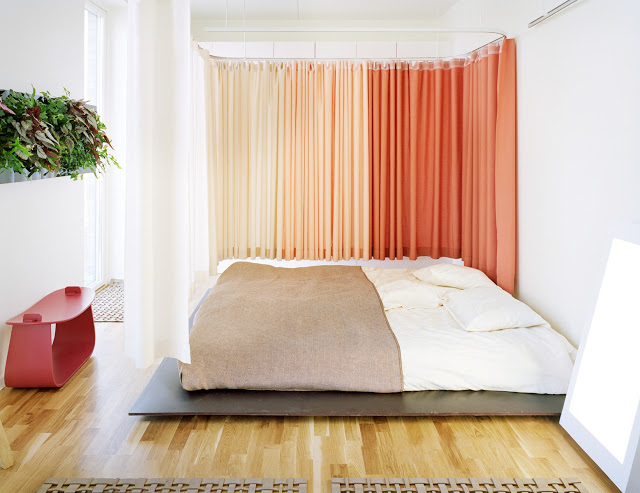 bedroom with orange floor length curtains, a low bed, light wood floor, and a red accent table
