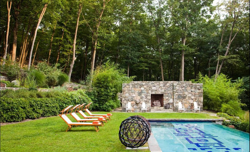Backyard with four lounge chairs, a pool with a graphic design on the bottom, grass, lots of trees and a stone fireplace.