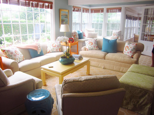 living room with paned windows with bamboo shades, two neutral sofas with blue and flower printed accent pillows, a yellow coffee table and three armchairs.
